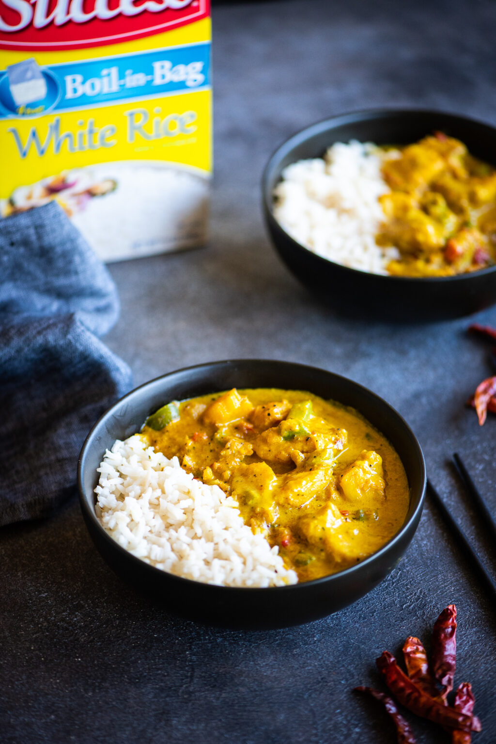 Two bowls of yellow fish curry with a box of success rice in the background