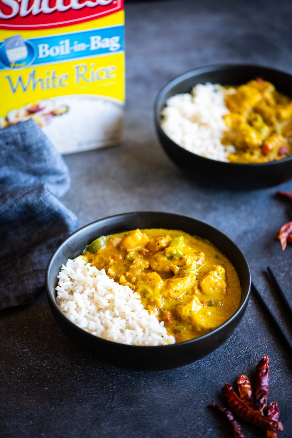 Two bowls of yellow fish curry with a box of success rice in the background