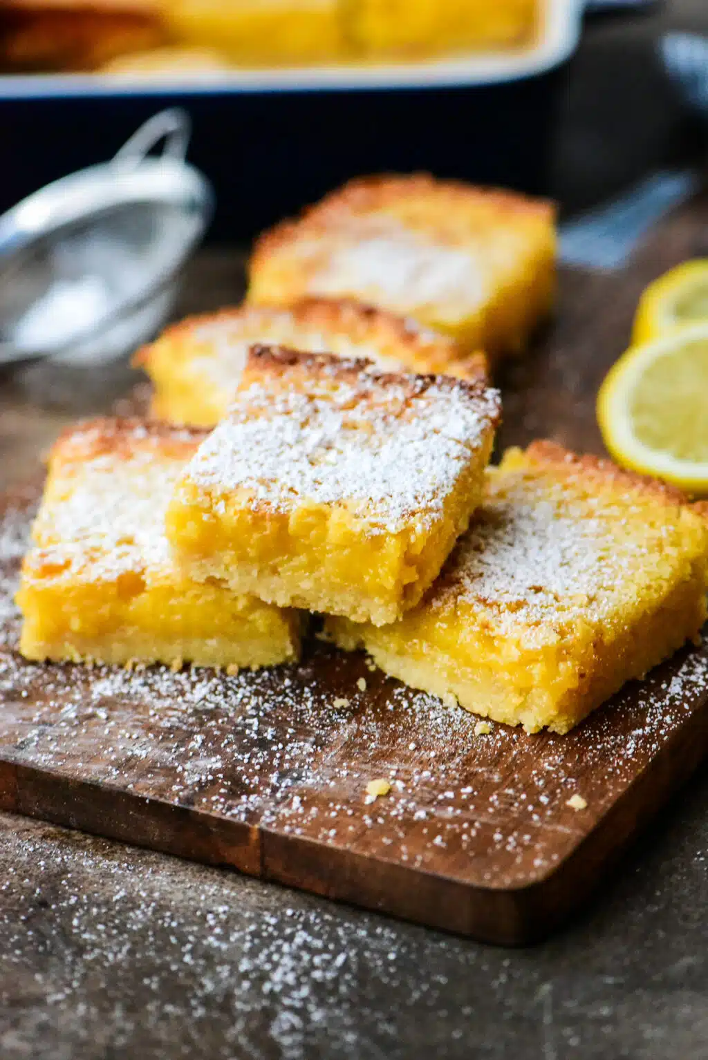 lemon bars stacked on a small wooden cutting board with slices of lemon